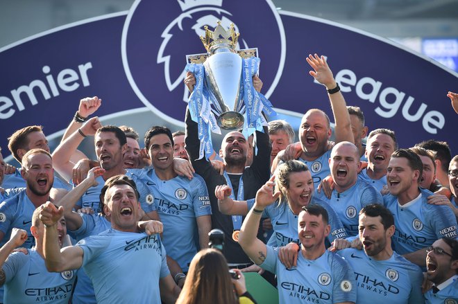 Strateg Manchester Cityja Pep Guardiola je 12. maja letos tako dvignil pokal za naslov zmagovalca premier league, obkrožen s svojimi nogometaši in njihovimi družinskimi člani. ManCity je prvi favorit za lovoriko tudi v sezoni, ki jo bodo odprli ta petek v Liverpoolu. FOTO: AFP