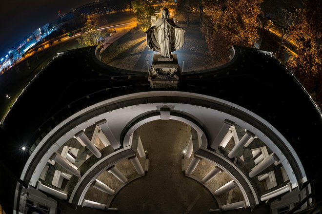 Slavolok Vrta vseh svetnikov, Žale, 2015 Marija Zavetnica s plaščem varuje umrle na notranji, vzhodni strani slavoloka in predstavlja svet mrtvih, mir in kontemplacijo. Nad njo se v hladni modri svetlobi dviguje stavba davčne uprave, zavetnice blaginje dr