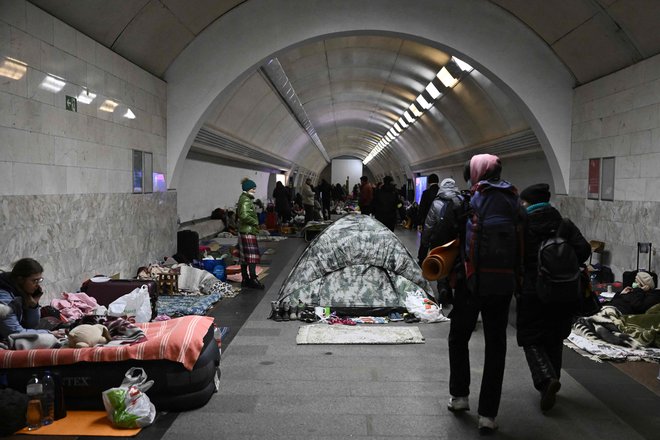 Civilisti hodijo mimo prebivalcev Ukrajine, ki uporabljajo podzemno postajo podzemne železnice kot zaklonišče pred bombnimi napadi. FOTO: Aris Messinis/AFP
