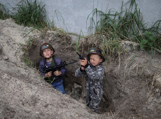 Maj 2022. Ukrajinska dečka, 12-letni Andrii in 6-letni Valentyn, stojita v jarku, ko se igrata ukrajinsko vojsko v bližini svojih hiš med rusko invazijo v vasi Stoianka v regiji Kijev. Foto: Gleb Garanich/Reuters
