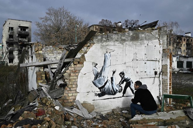 Ulični umetnik Banksy je na svojem uradnem profilu na Instagramu objavil posnetek najnovejšega dela, ki ga je ustvaril na pročelju poškodovane stavbe v Ukrajini. V Kijevu je upodobljen otrok, ki v judoističnem dvoboju zavihti odraslega moškega, ki spominja na Vladimirja Putina, za katerega je znano, da ima črni pas v judu. Foto: Genya Savilov/Afp
