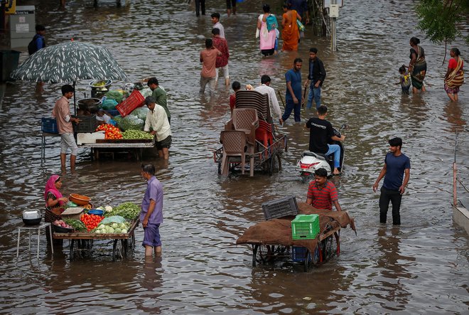 Indijci kupujejo zelenjavo pri uličnih prodajalcih, medtem ko se drugi prebijajo skozi poplavljeno cesto po močnem deževju v Ahmedabadu. Foto: Amit Dave/Reuters