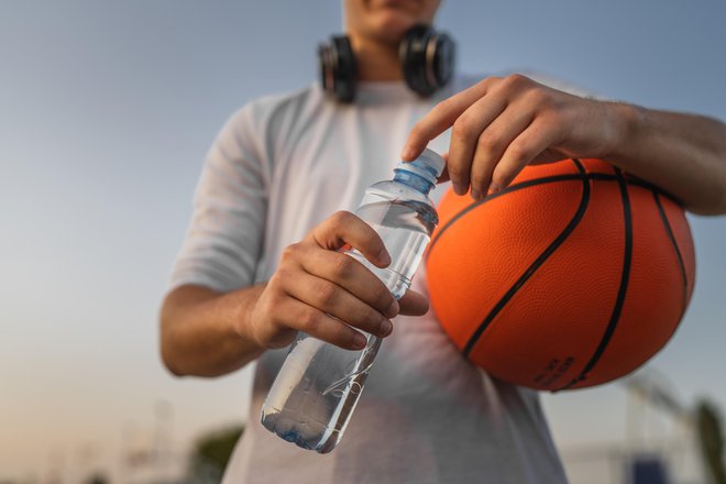 close up on hands midsection of unknown caucasian man teenager open plastic bottle of water outdoor in sunny day doing sports with basketball