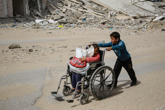 Deček potiska deklico na invalidskem vozičku mimo porušene stavbe v Gazi. Dostave nujno potrebne dobrodelne pomoči v glavnem ovira Izrael, ki ga je Meddržavno sodišče (ICJ) v četrtek pozvalo, da mora brez odlašanja zagotoviti nujno pomoč prebivalcem Gaze. Ob tem je opozorilo na hudo lakoto v palestinski enklavi, ki je že skoraj pol leta tarča nenehnih izraelskih napadov. Foto: Afp