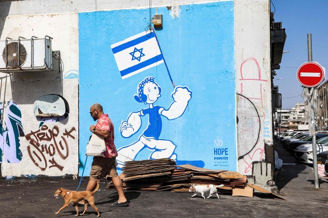 Moški sprehaja pse mimo grafita v Tel Avivu, na katerem je upodobljen lik Olive Oyl iz risanke Popeye z mišičastimi rokami in izraelsko zastavo. Foto: Gil Cohen-magen/Afp